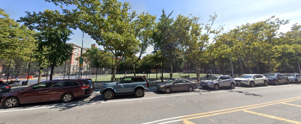 A street view of a park with a fence, trees, and parked cars along the curb.